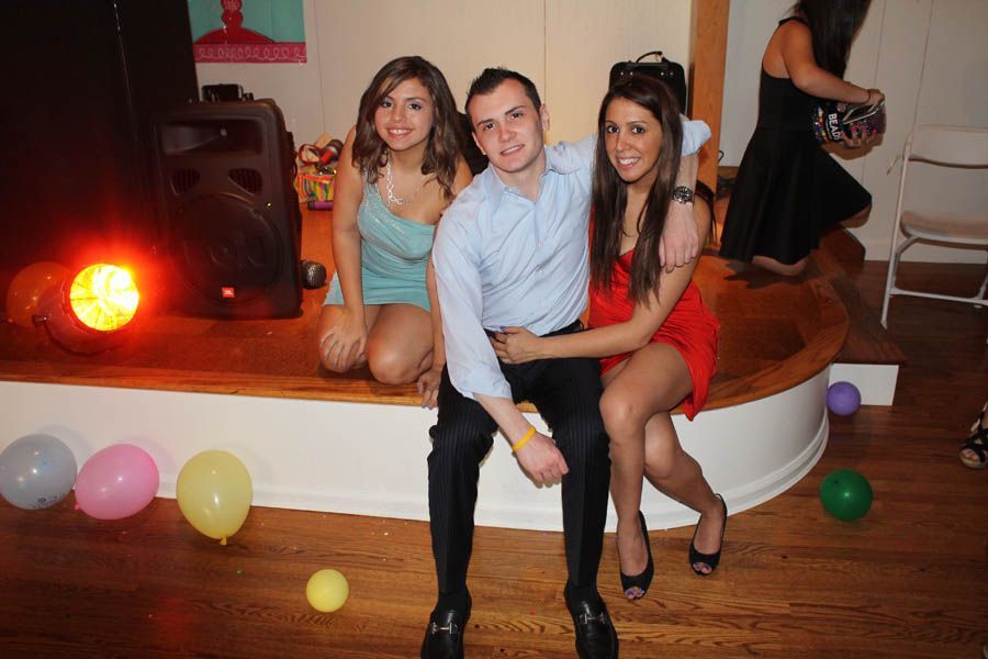 Man flanked by two women, all seated on a stage, at a party. Balloons on floor, stage lit with a spotlight.
