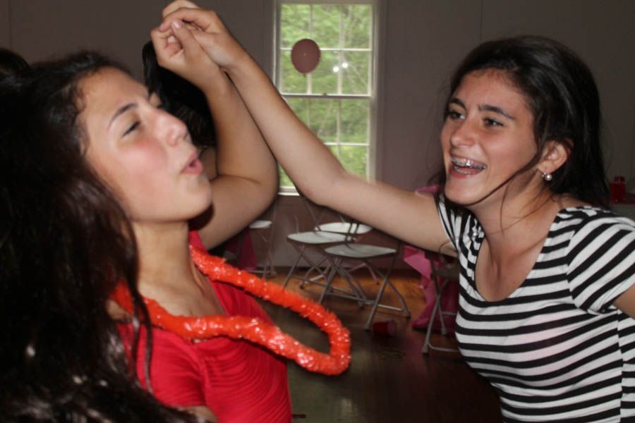 Two girls, one with braces, play with a lei indoors, both smiling and interacting.