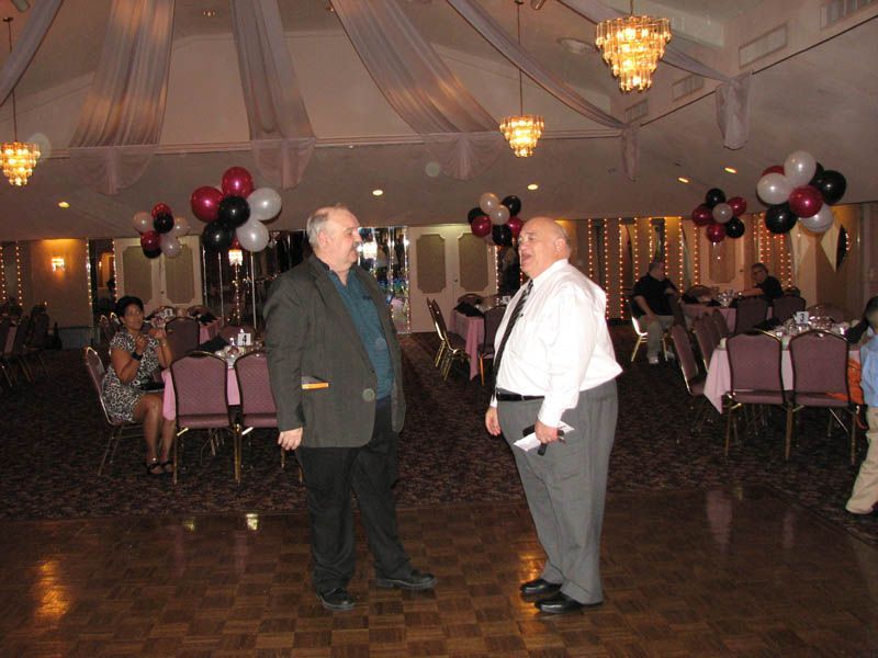 Two men conversing on a dance floor in a decorated banquet hall, surrounded by tables and balloons.