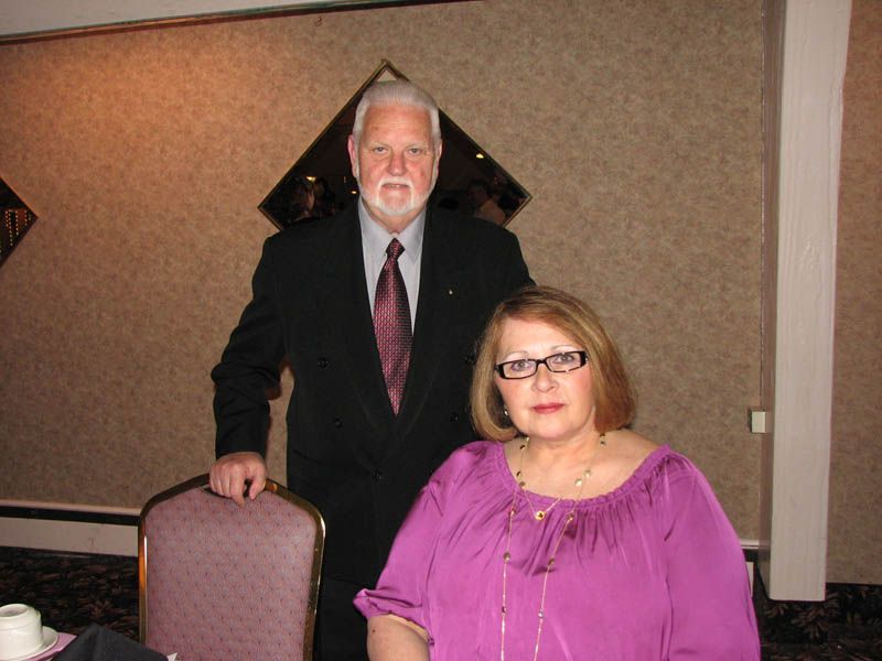 Man in suit stands behind woman in purple top, both in a formal setting.