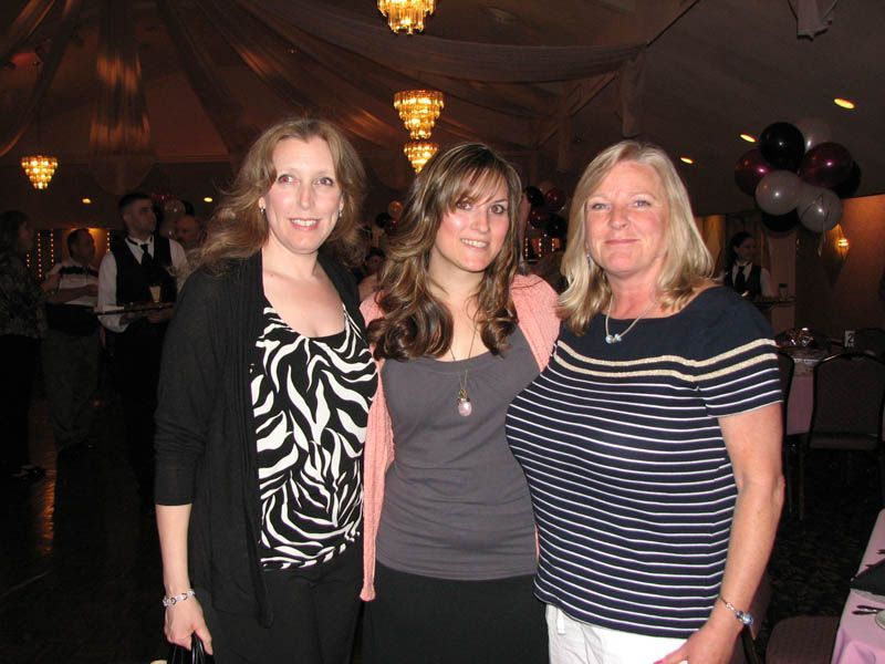 Three women smiling indoors, posing together. One in black and white print, another in pink, and one in stripes.