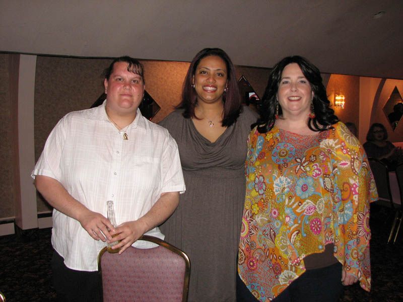 Three people smiling, posed inside. One wears a floral top, one a brown dress, and one a white shirt.