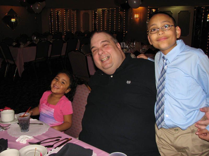 Man smiling with a boy and girl at a table in a dimly lit banquet hall.