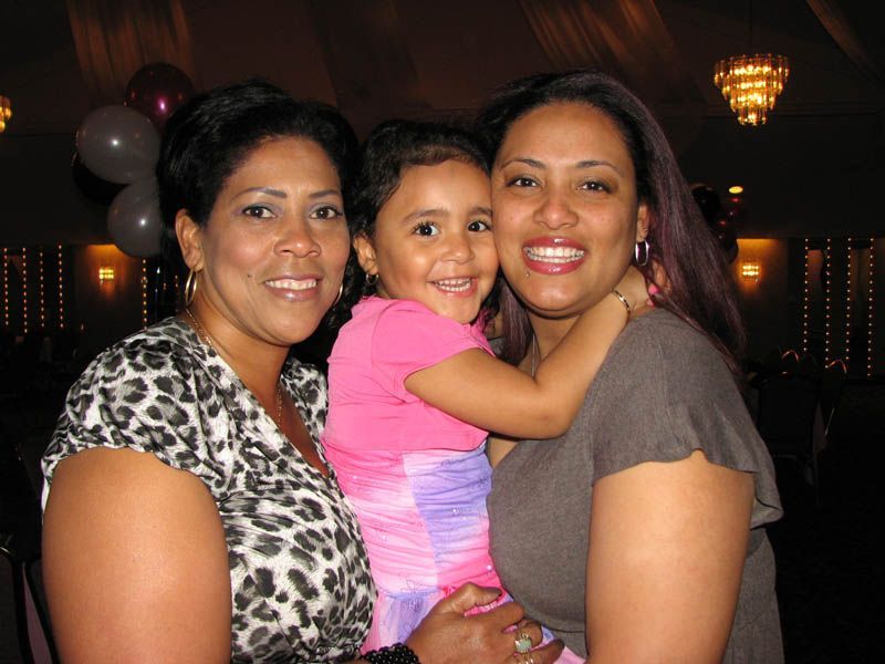 Two women and a child smile, embracing indoors; balloons and lights in the background.