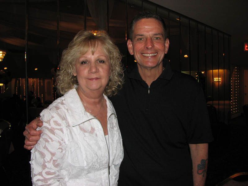 Woman and man smiling, posing indoors. Woman wears white shirt, man wears black. Dark background.