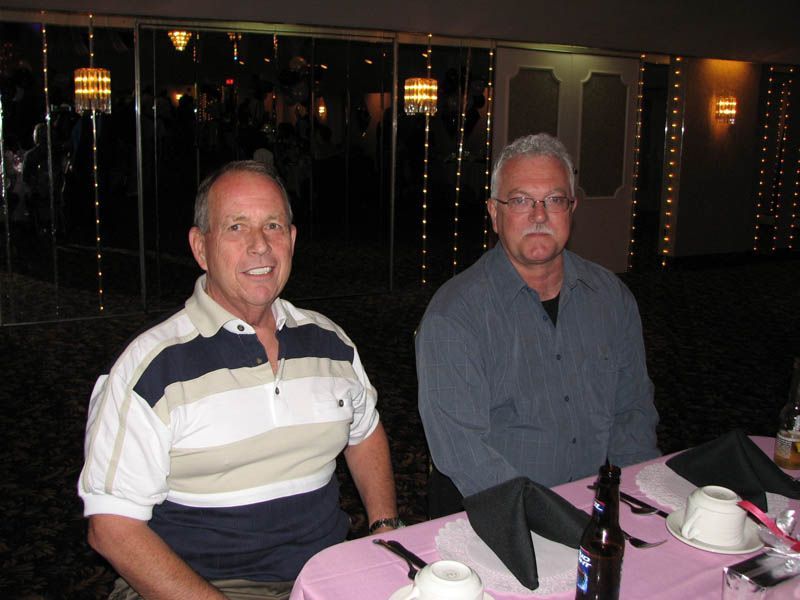 Two men seated at a table, one smiling, the other with a neutral expression, in a banquet hall.