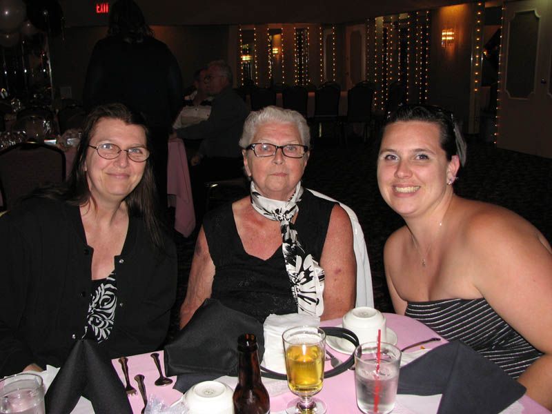 Three women seated at a table, smiling. One in black, white scarf; others in black and a striped top. Dimly lit room.