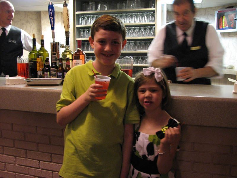 Two children at a bar, holding red drinks, smiling. Bartenders behind them.