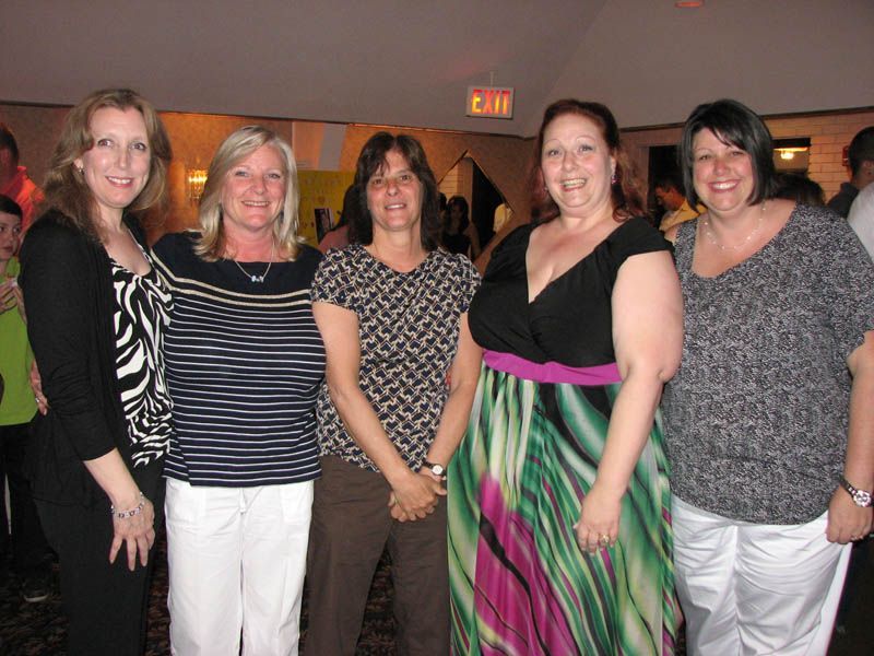 Five women smiling, posing for a photo indoors, wearing various patterned tops, dresses, and pants.