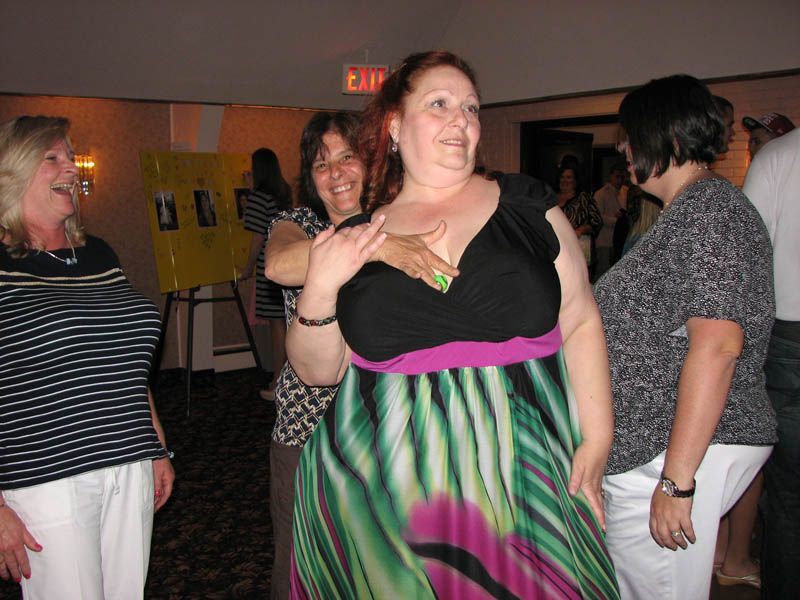 Four women in a room, two touching the central woman wearing a colorful dress. They're all smiling in a social setting.