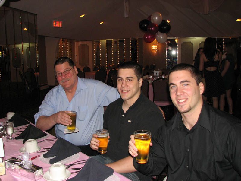 Three men at a table, toasting with beer. Formal setting with pink tablecloth, balloons, and other party-goers.