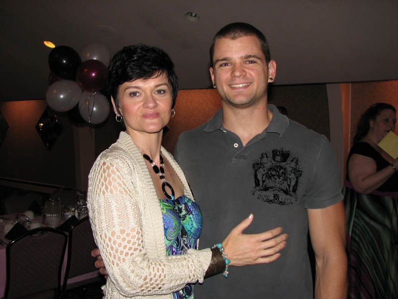 Woman and man pose together in a room with balloons; woman wears a sweater and dress; man wears a polo.