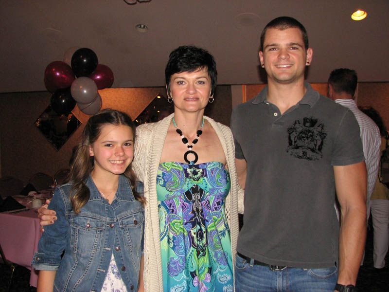 Woman in colorful dress poses with a girl in denim jacket and a man in grey polo shirt; balloons in background.