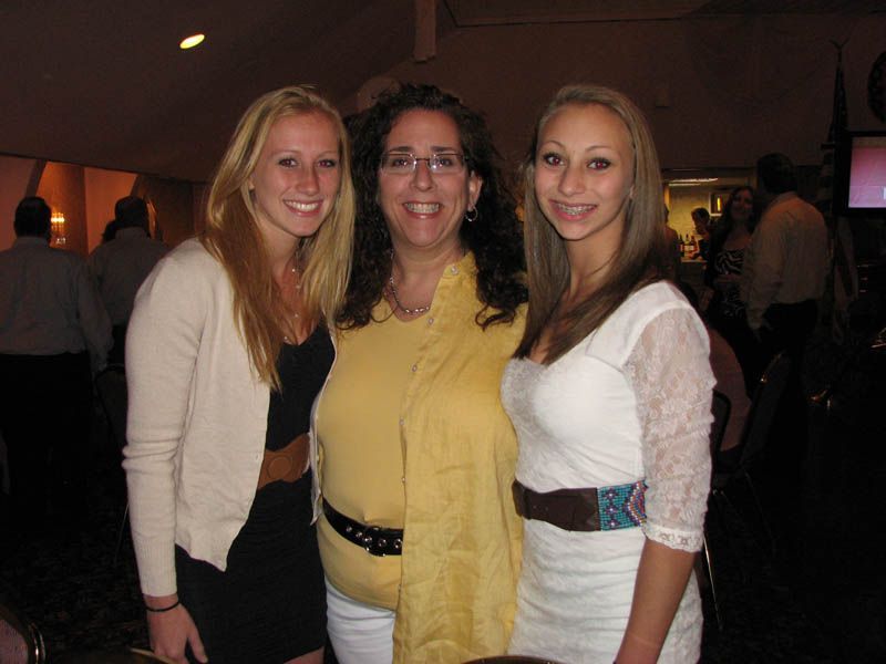 Three people smiling, posing for photo indoors. Woman in yellow with two younger women in black and white outfits.
