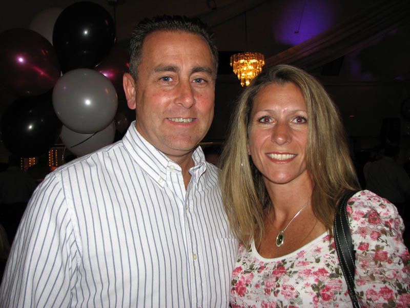 Man and woman smiling, indoors. Man in striped shirt, woman in floral top. Balloons and chandelier visible.