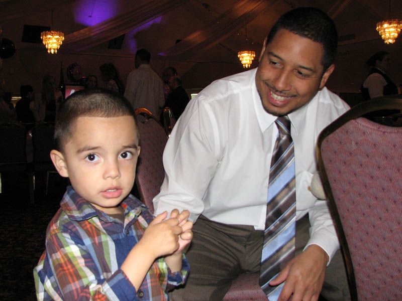Man in a white shirt and tie smiles, holding the hand of a child in a plaid shirt. They are in a well-lit indoor space.