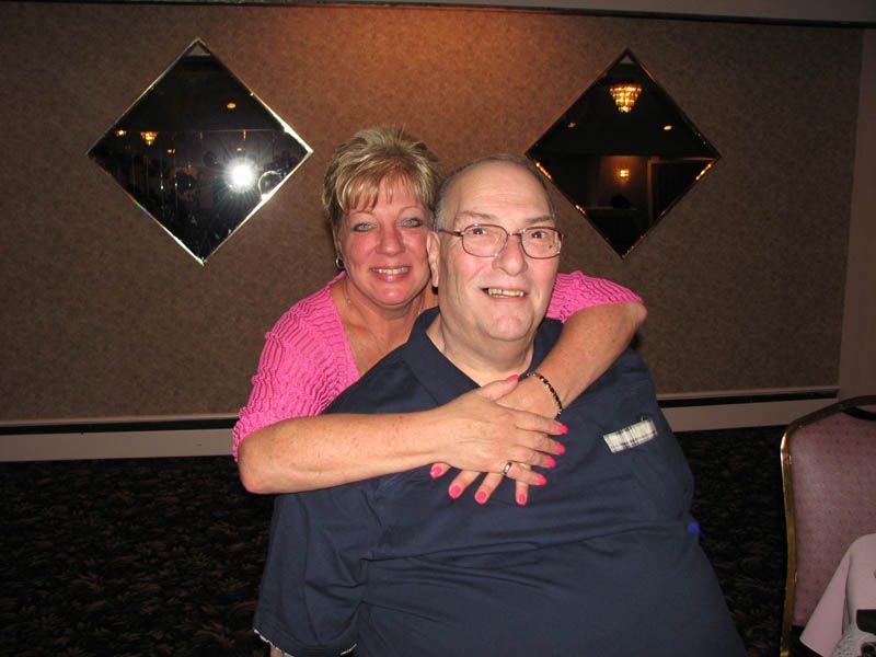 Woman hugs man; both smiling. Woman wears pink sweater, man a blue shirt. Background has diamond mirrors.