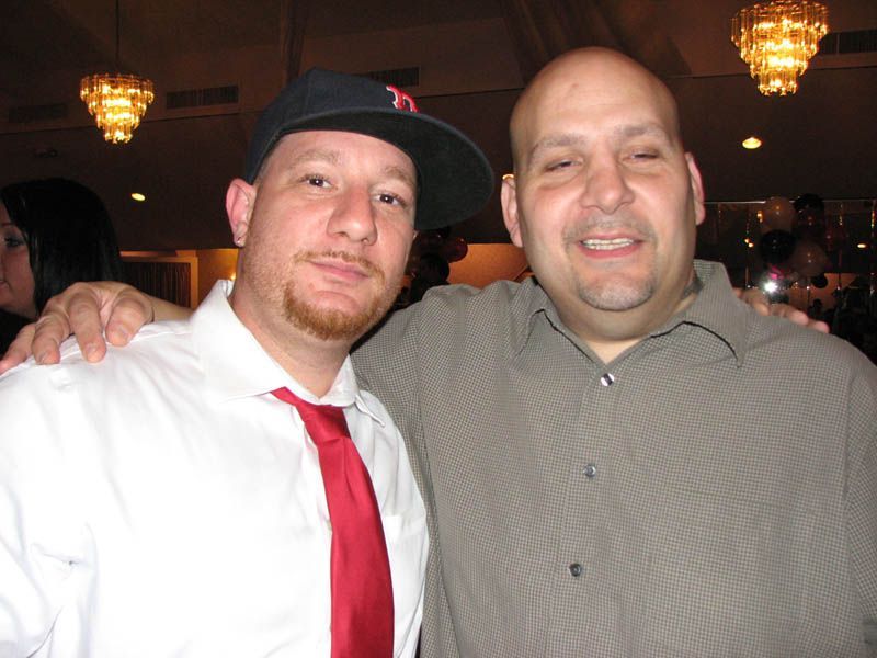 Two men pose together. One in a red tie and a backward cap, the other with a hand on his shoulder, both smiling in a room.