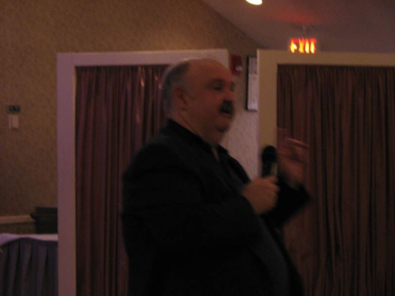 Man in dark suit speaking into a microphone, standing in front of a curtained backdrop.