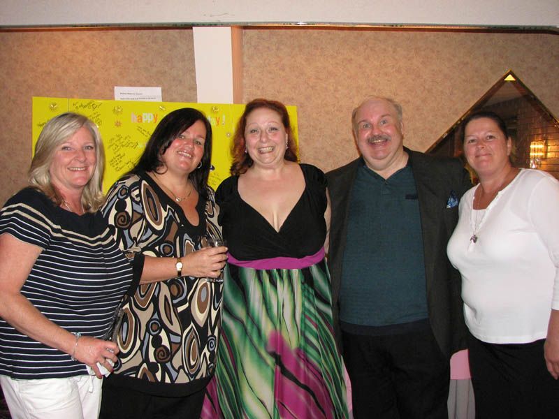 Five people smiling together indoors; the central woman wears a colorful dress.