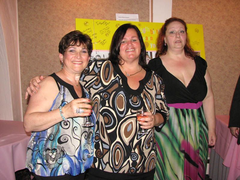 Three women smiling, holding drinks at an event; one has her arm around another. Background includes a table and a sign.
