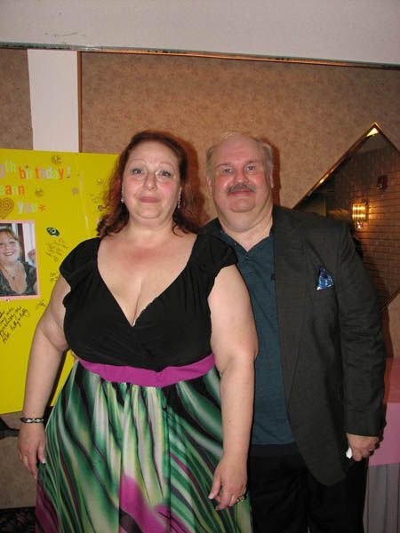 Woman in black top and colorful dress, stands with man in a blazer, both smiling. Background has a poster.