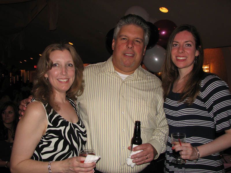 Three people smiling at a party, man in striped shirt holds a bottle, women hold drinks. Balloons in the background.