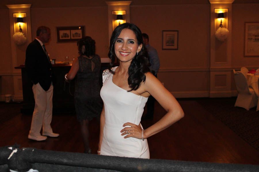 Woman in white dress smiles, poses at an event. Dark hair, hand on hip. Event setting with others in background.