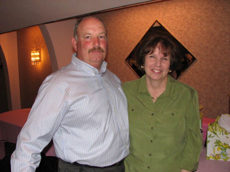 Man in blue striped shirt and woman in green shirt smiling, standing indoors, near a table.