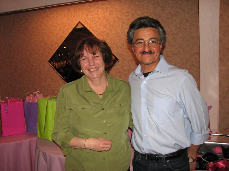 Two people smiling, posing together indoors near gifts. The woman in green, the man in a blue shirt.
