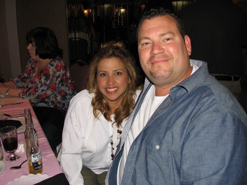 Smiling couple at a table, woman in white shirt, man in denim. Indoor setting with a woman in the background.