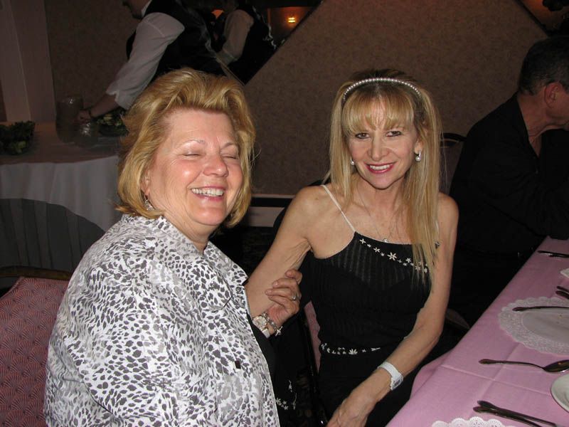 Two women smiling, seated at a table. One in a black dress, holding the other woman's arm. Party setting.