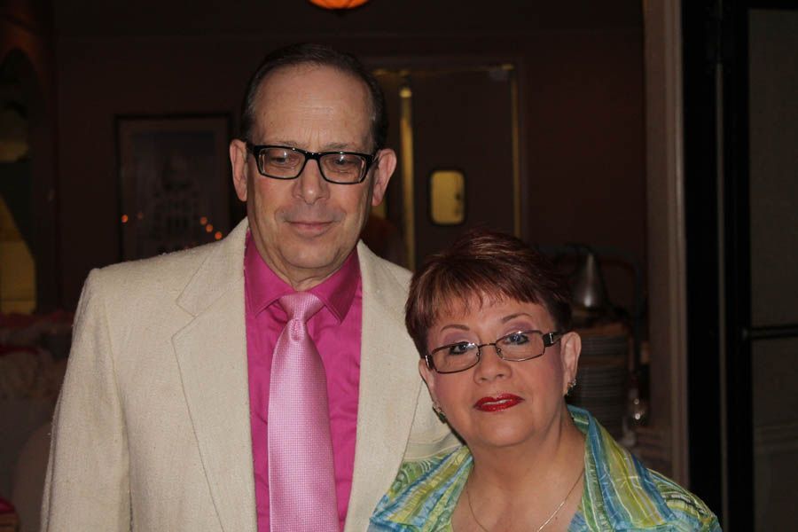 Man and woman smiling, indoors. Man wears white blazer and pink tie. Woman wears patterned jacket and glasses.