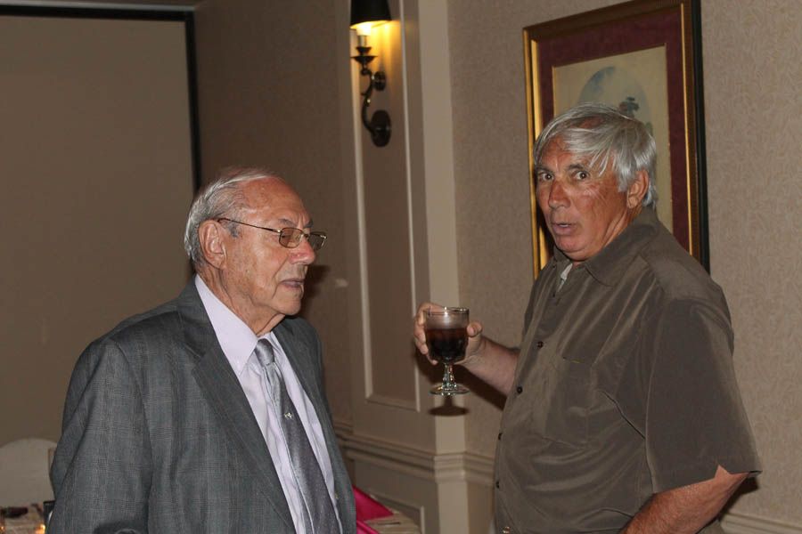 Two older men talking; one holds a drink. Formal setting, light brown and white walls.