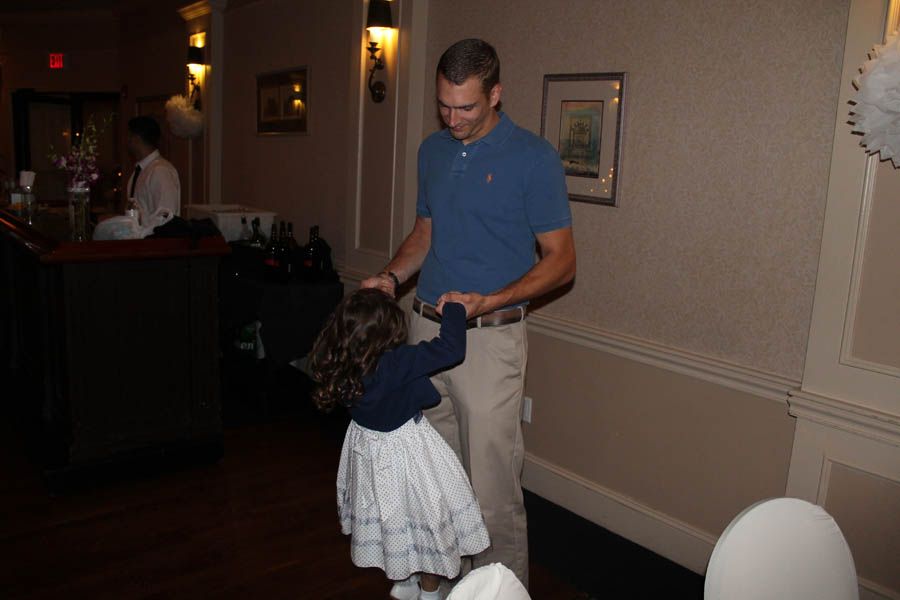 Man in blue shirt twirling a small girl in a white dress indoors, smiling.