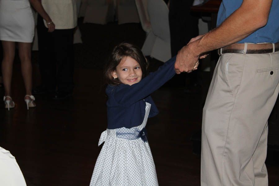 Girl dances, holding a man's hand. She wears a white dress and blue jacket; smiles. Dark interior with others.