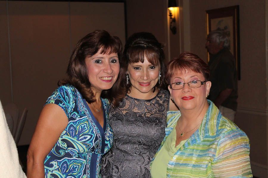 Three women pose together indoors, smiling. One in blue print, one in gray dress, one with glasses.