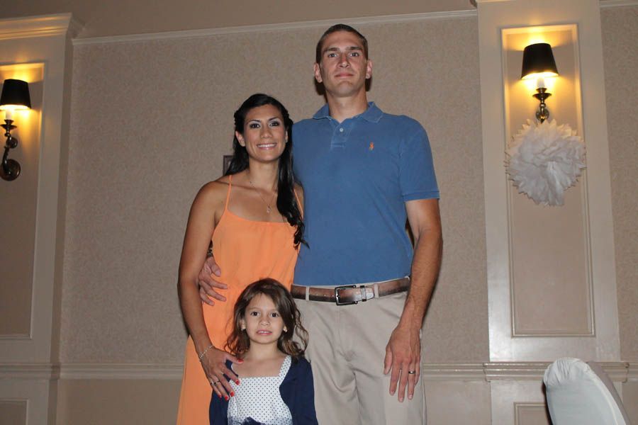 Family of three poses indoors. Woman in orange dress, girl in blue, tall man in blue shirt, beige walls.