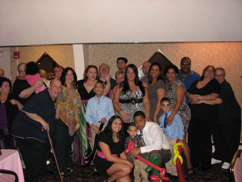 Large group of people smiling at an indoor event, some children. Pink table cloth.