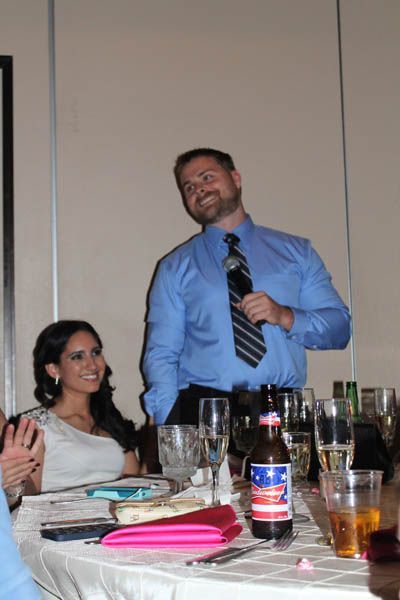 Man in blue shirt, holding a microphone, giving a speech at a table. Woman in white dress smiles, next to him.