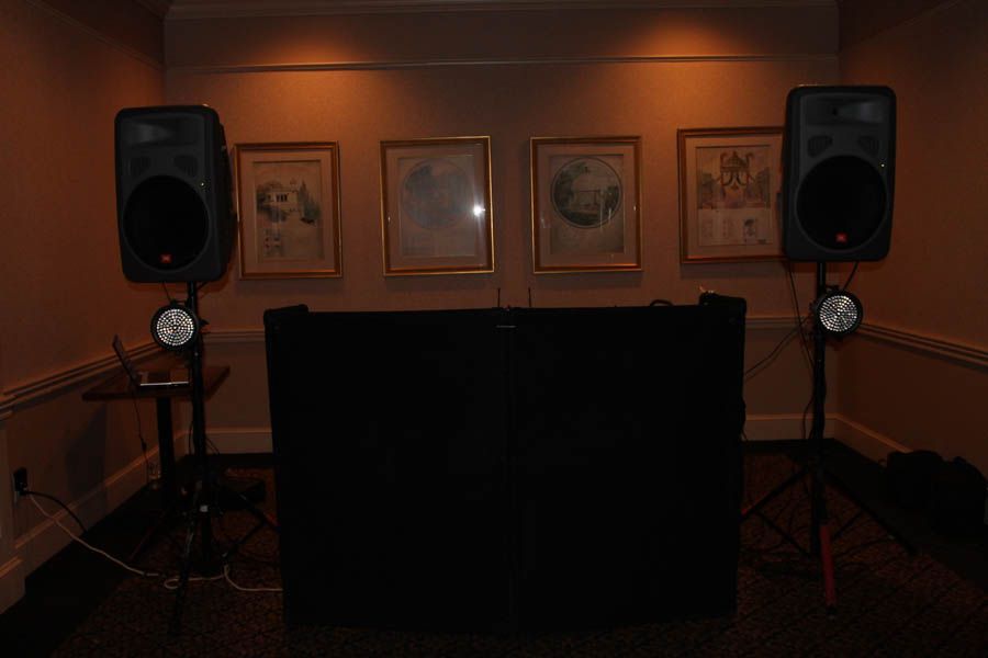 DJ setup: two speakers on stands, black facade, and stage lights against a wall with framed art.