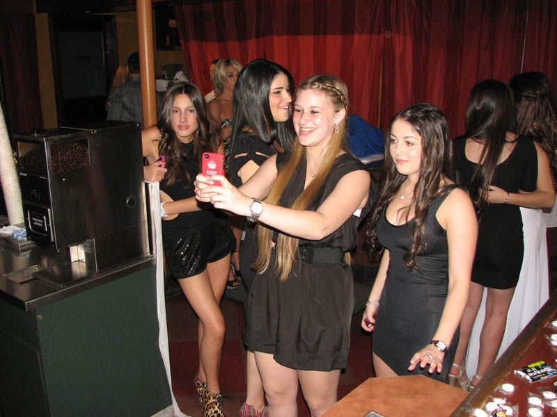 Five young women in black outfits pose for a selfie inside a dimly lit venue with red curtains.