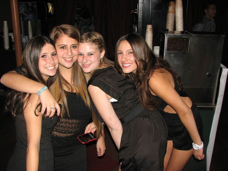 Four smiling women in black dresses pose together in a dimly lit space, arm in arm.