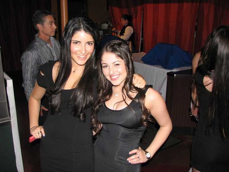 Two women in black dresses pose together indoors. One has arm around the other; both smile.