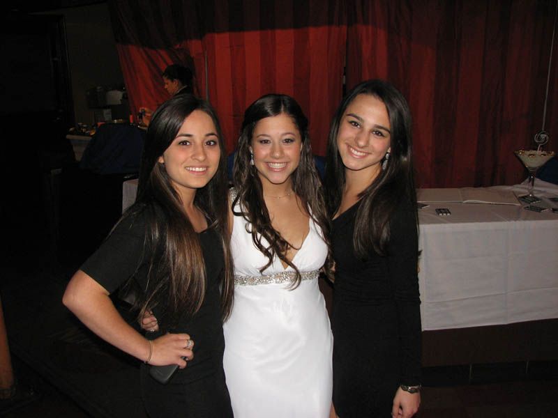 Three smiling women, two in black dresses flanking one in a white gown, pose at an event.