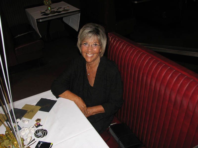Woman in black top smiles, seated in a red booth at a table, low lighting.