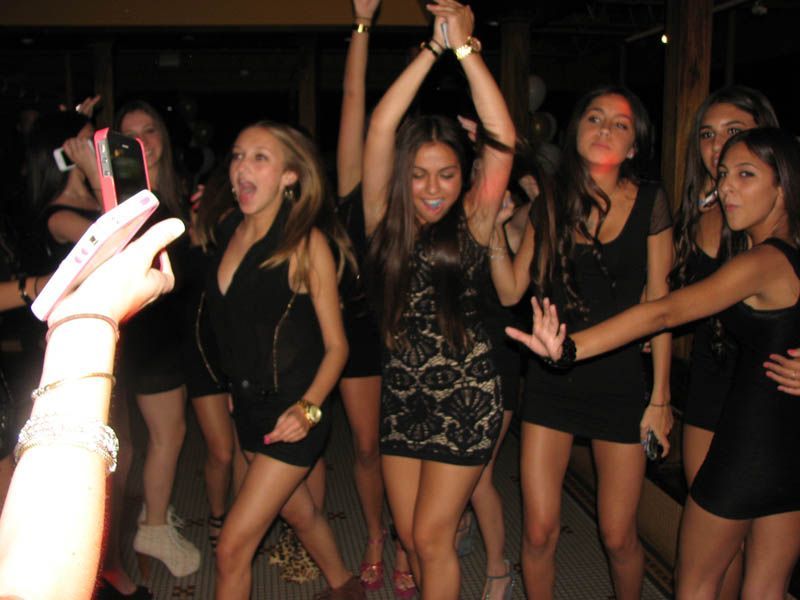 Group of young people dancing at a party, many in black dresses, some with arms raised, lit by dim lights.