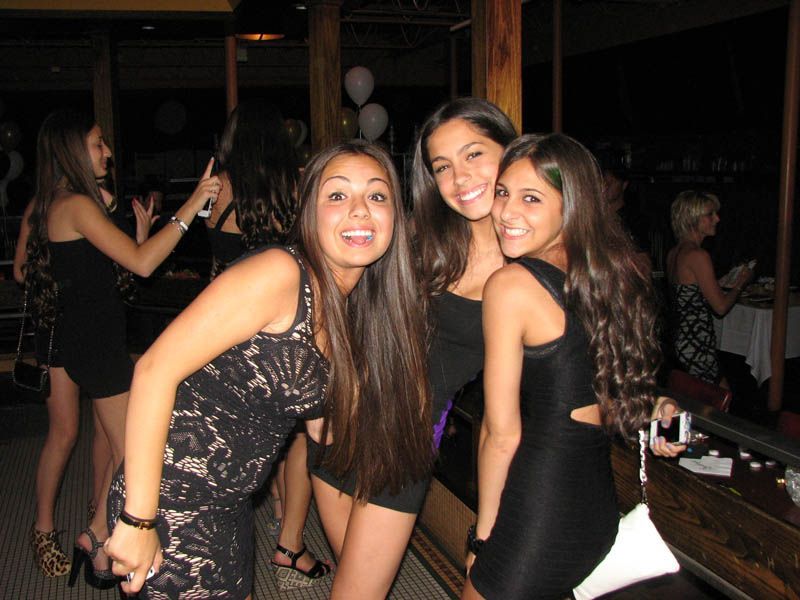 Three young women smiling, in black dresses, at a party, with other people in the background.