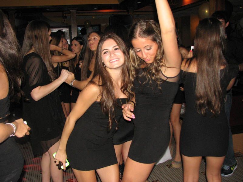 Women in black dresses dancing at a club, smiling, with arms raised.