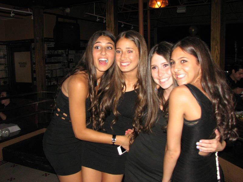 Four young women smiling and posing together in a dimly lit bar, wearing black dresses.
